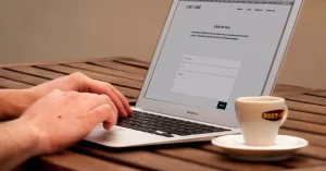 Close-up of hands typing on a laptop next to a cup of coffee on a wooden desk.